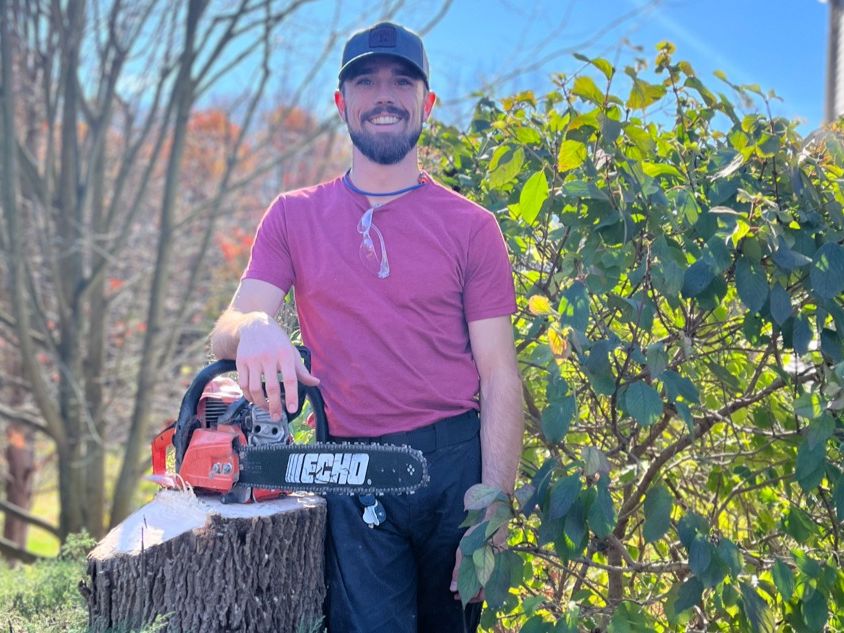 A smiling man in a red shirt and dark cap leans on a chainsaw resting on a tree stump, with a leafy shrub and bare trees behind him.